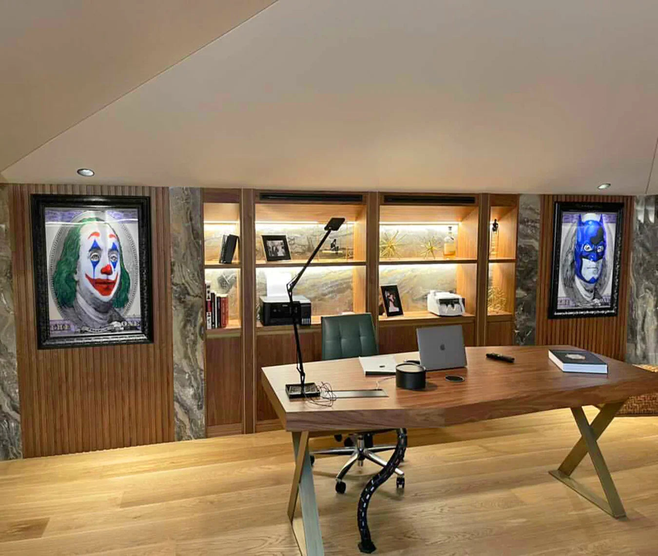 Modern office desk with laptop, lamp, and bookshelf flanked by Joker and Batman framed art