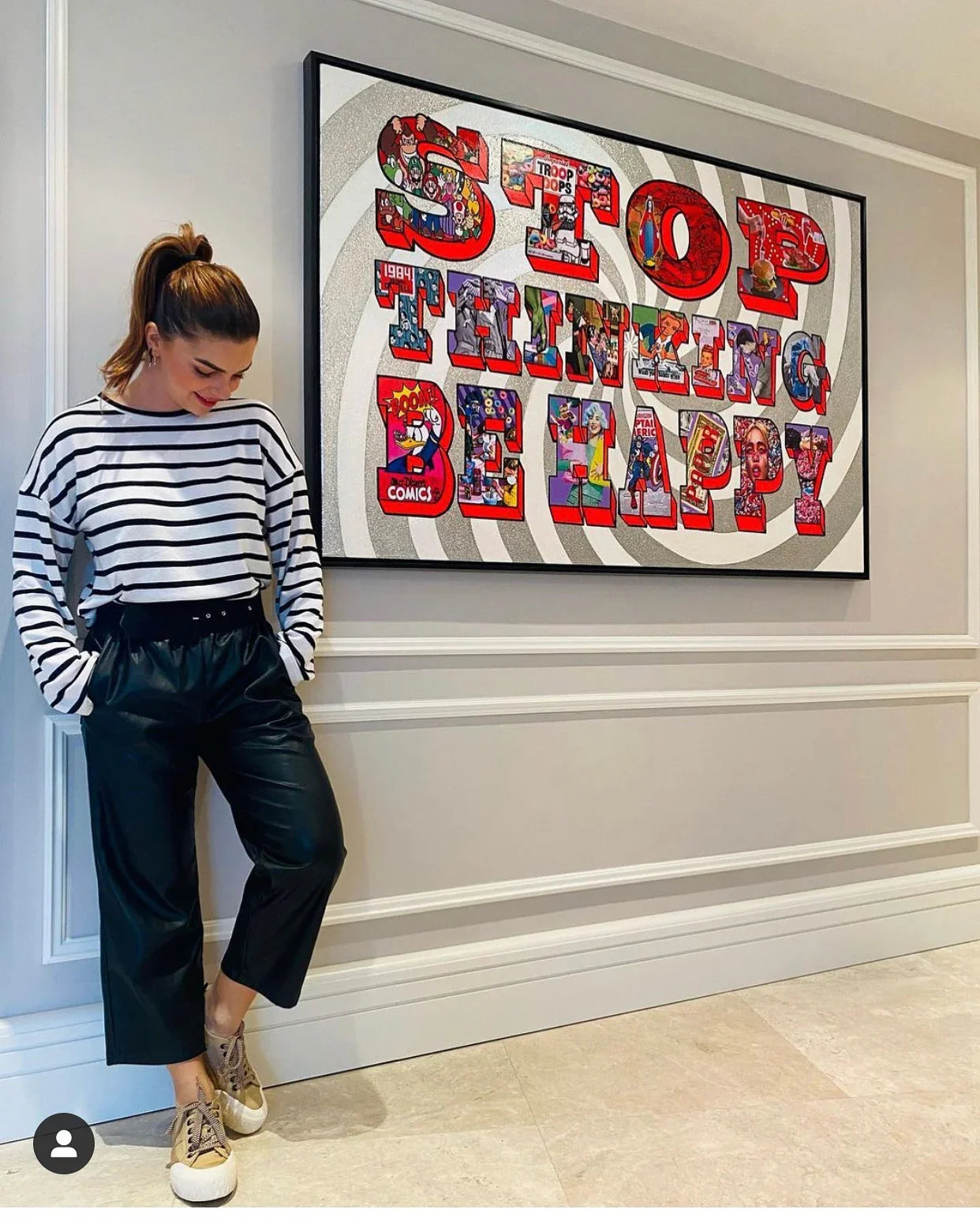 Woman in striped shirt and black pants standing next to colorful 'Stop Thinking Be Happy' wall art